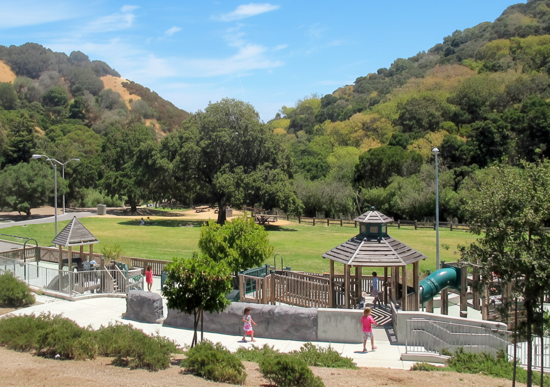 Playground and green space at Laurelwood Park in San Mateo on a sunny day, with children playing near a gazebo and slide, surrounded by trees and hills.