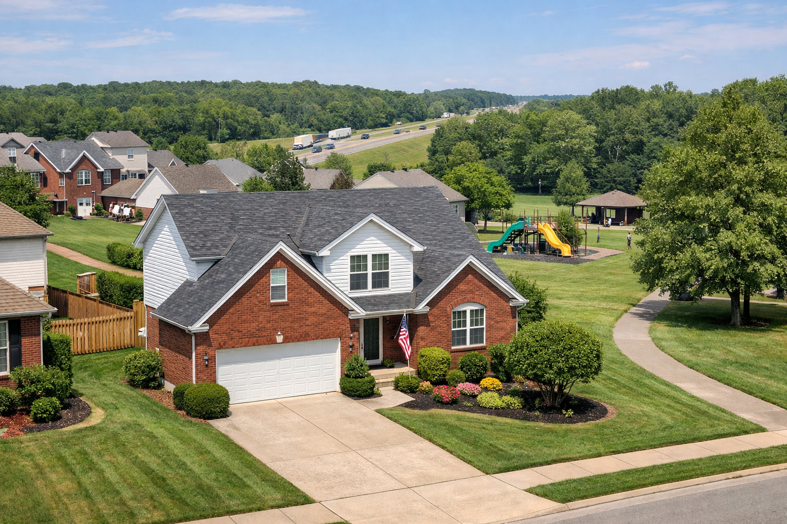 A bright daytime photo of a red brick and white siding two-story home in Fern Creek, Kentucky, featuring a two-car garage, manicured lawn, landscaped shrubs, and a small American flag by the front door. In the background, there is a neighborhood park with a playground, mature trees, and the Gene Snyder Freeway visible beyond the homes under a clear blue sky.