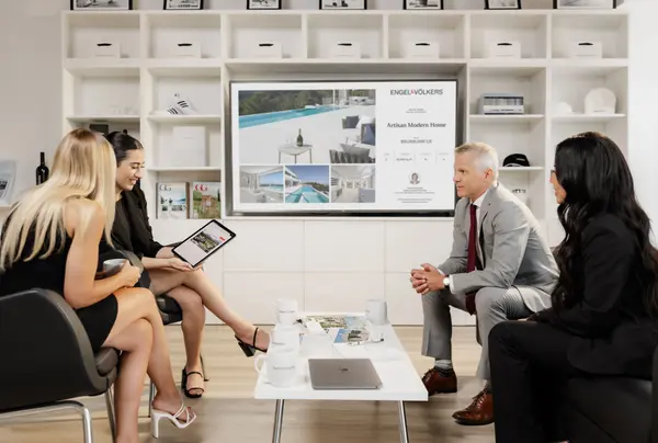 Engel & Voelkers real estate agents and customers in a real estate office with shop TV on the wall reviewing listings on a tablet