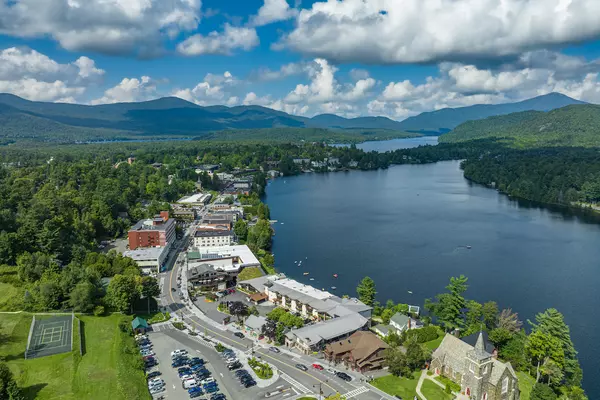 Aerial view of downtown Lake Placid, NY real estate and shops along Mirror Lake, with Adirondack Mountains in the background on a sunny summer day.
