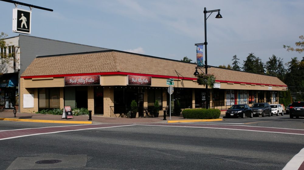 Single-storey strip of shops with Brick Alley Bistro signs beside a crosswalk, and a street sign reading Fraser Hwy and 271 St