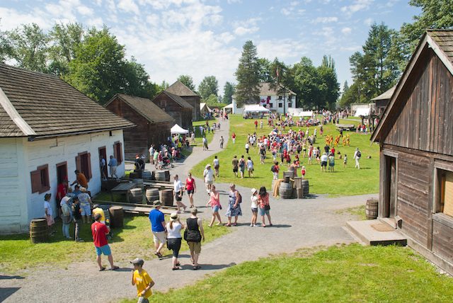 Visitors walking a gravel path between a white-walled building and a log building with wooden barrels, and a crowd gathered on the lawn beyond, at a reconstructed fort on a sunny day
