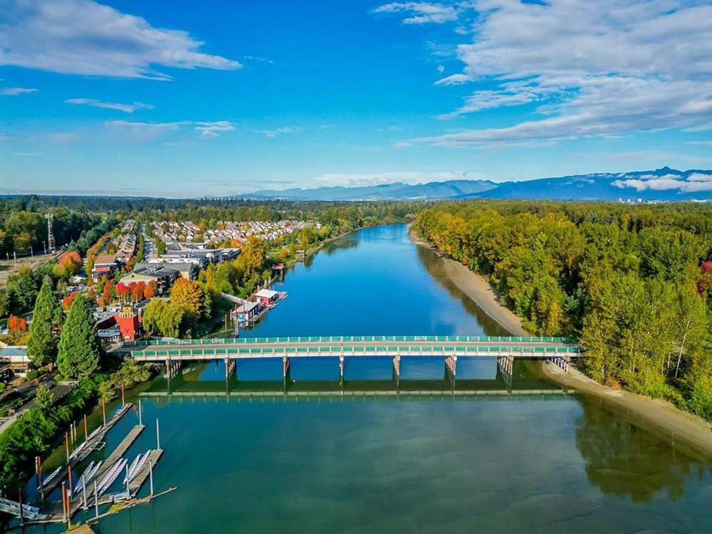 Aerial view in autumn of homes and low-rise buildings along the Fraser River, with a long green-railed bridge crossing the water, boat docks in the foreground and mountains on the horizon