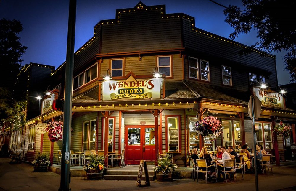 Wendel's Books and Fresh Food Cafe at dusk, a wooden two-storey storefront outlined in string lights, with hanging flower baskets and people dining at patio tables out front