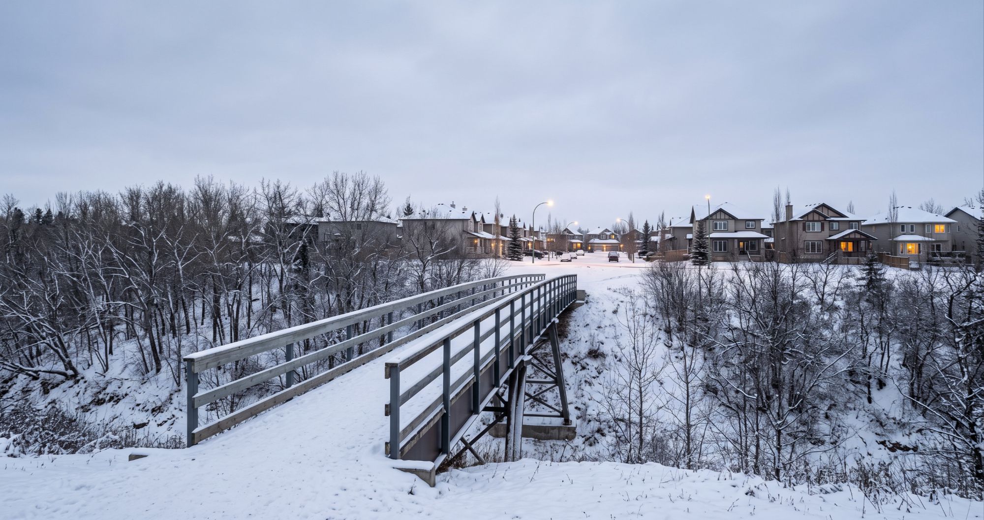 A snow-covered pedestrian bridge in Edmonton connects to a residential street of suburban homes, representing the transition of bridge financing between two properties