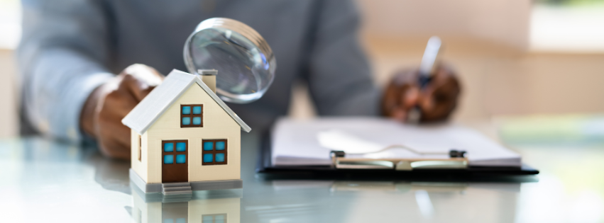 Close-up of a miniature house model with a magnifying glass, symbolizing a home inspection, with a professional reviewing documents in the background.
