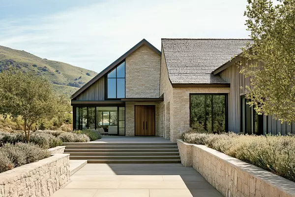 A modern farmhouse with a gable roof, located in the rolling hills of California's wine country, features natural stone walls and large windows that frame the vineyards