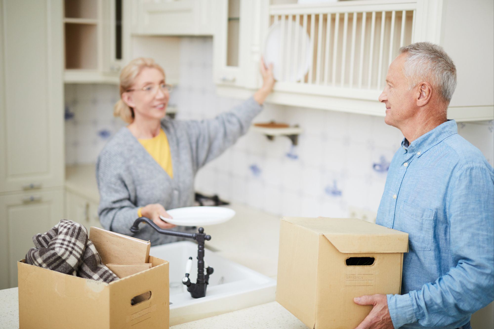 Grey-haired man with carton box standing by sink in the kitchen and talking to his wife during relocation