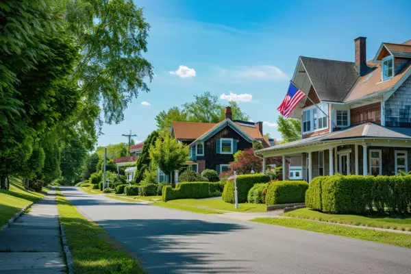 American flags flying in front of a beautiful home on a quaint street with green hedges and trees on a sunny day in a small town