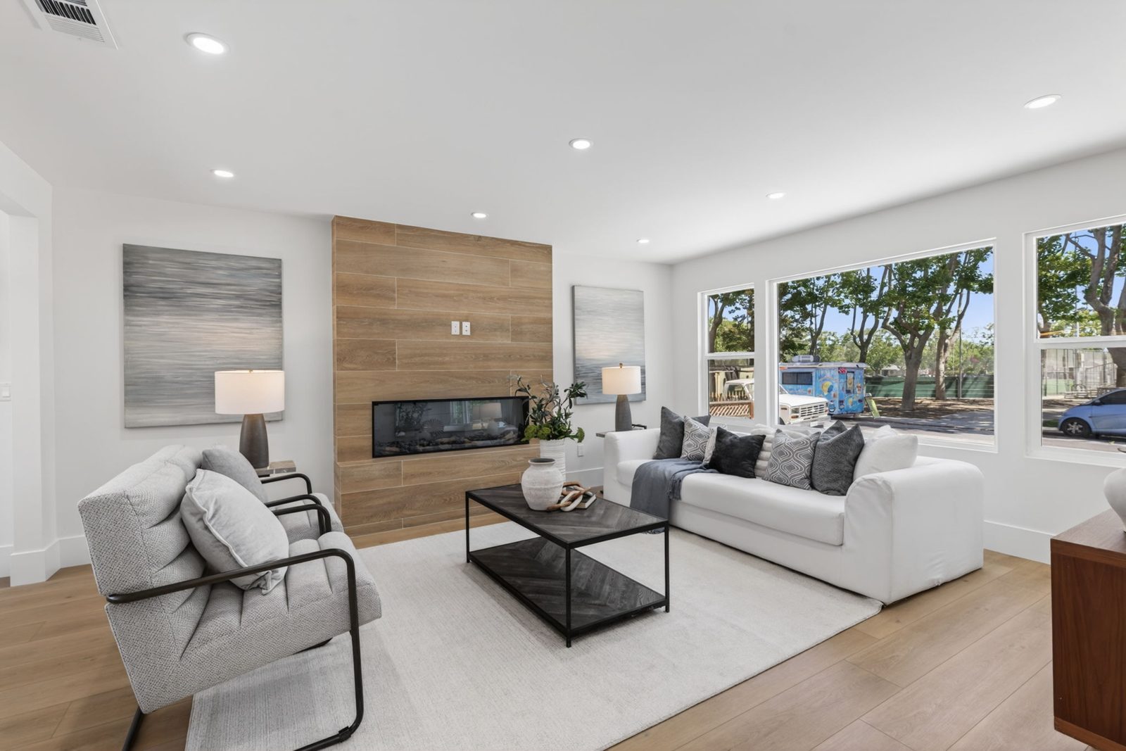 Living room with wide plank floors, a wood paneled fireplace wall and big new windows.