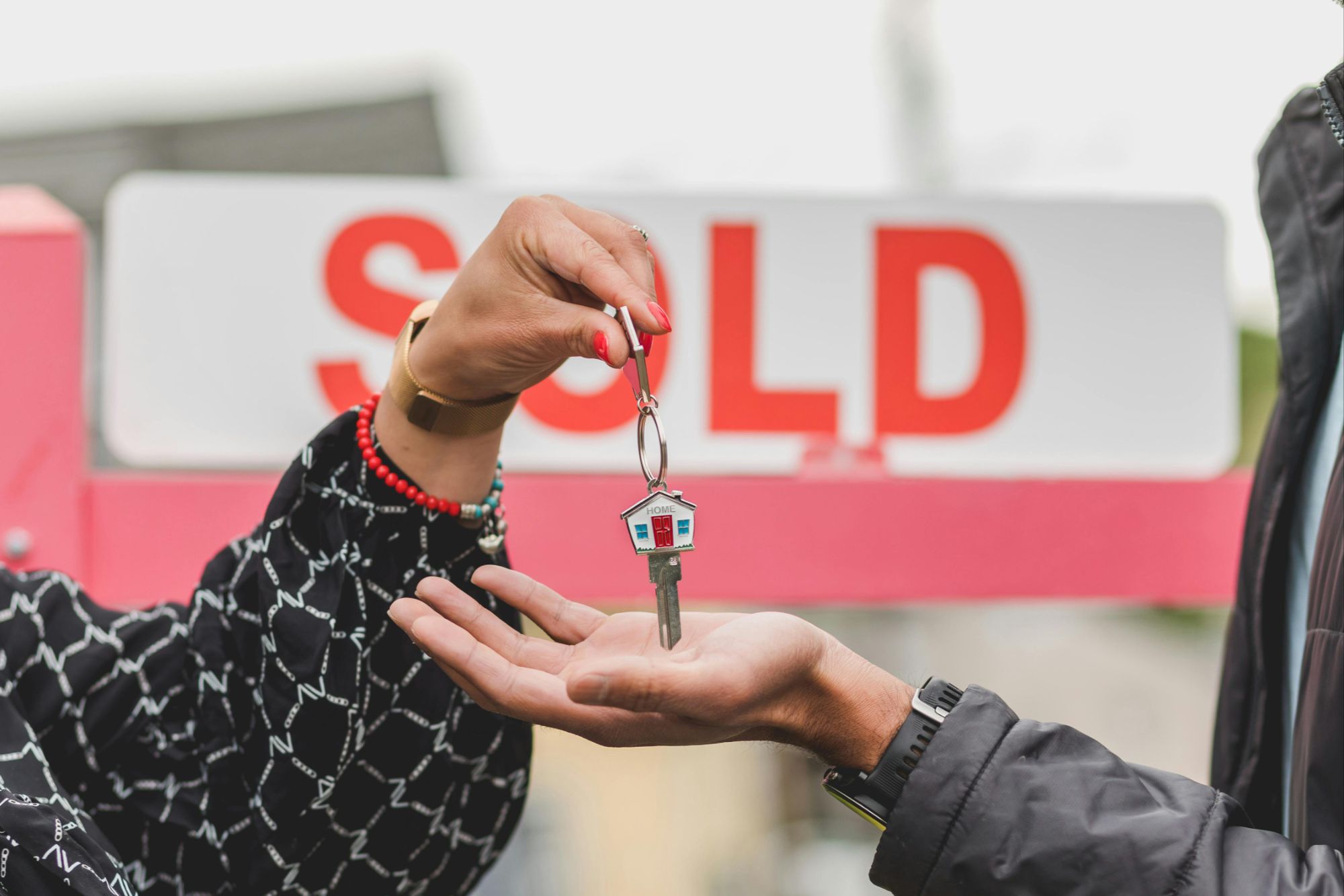 Close-up of a Boulder homeowner receiving house keys with a house-shaped keychain in front of a blurred "SOLD" sign, symbolizing a successful real estate closing or mortgage buyout.