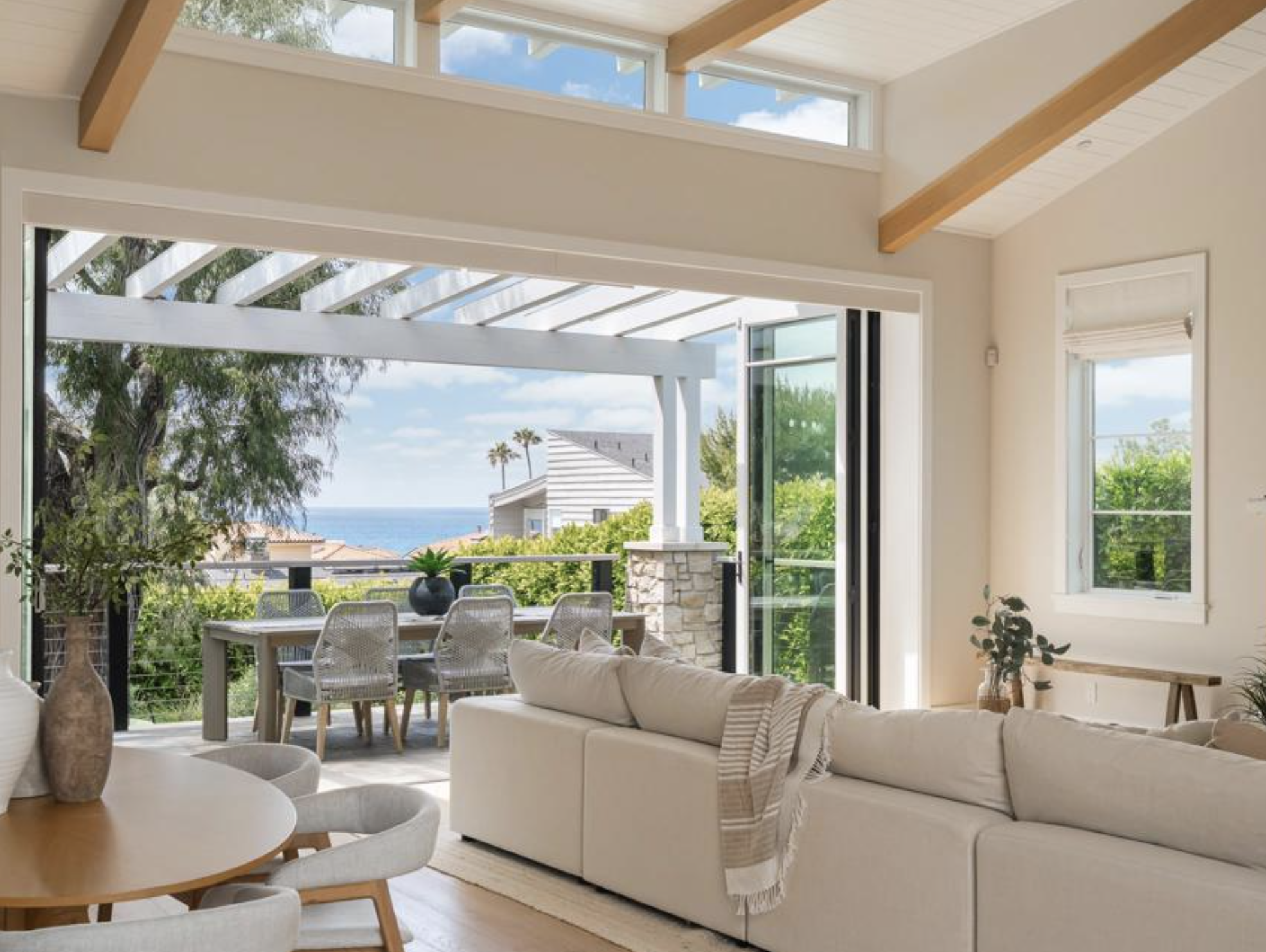  Luxury open-concept living room featuring a cream sectional sofa, vaulted wood ceilings, and retractable glass doors opening to Pacific Ocean views.