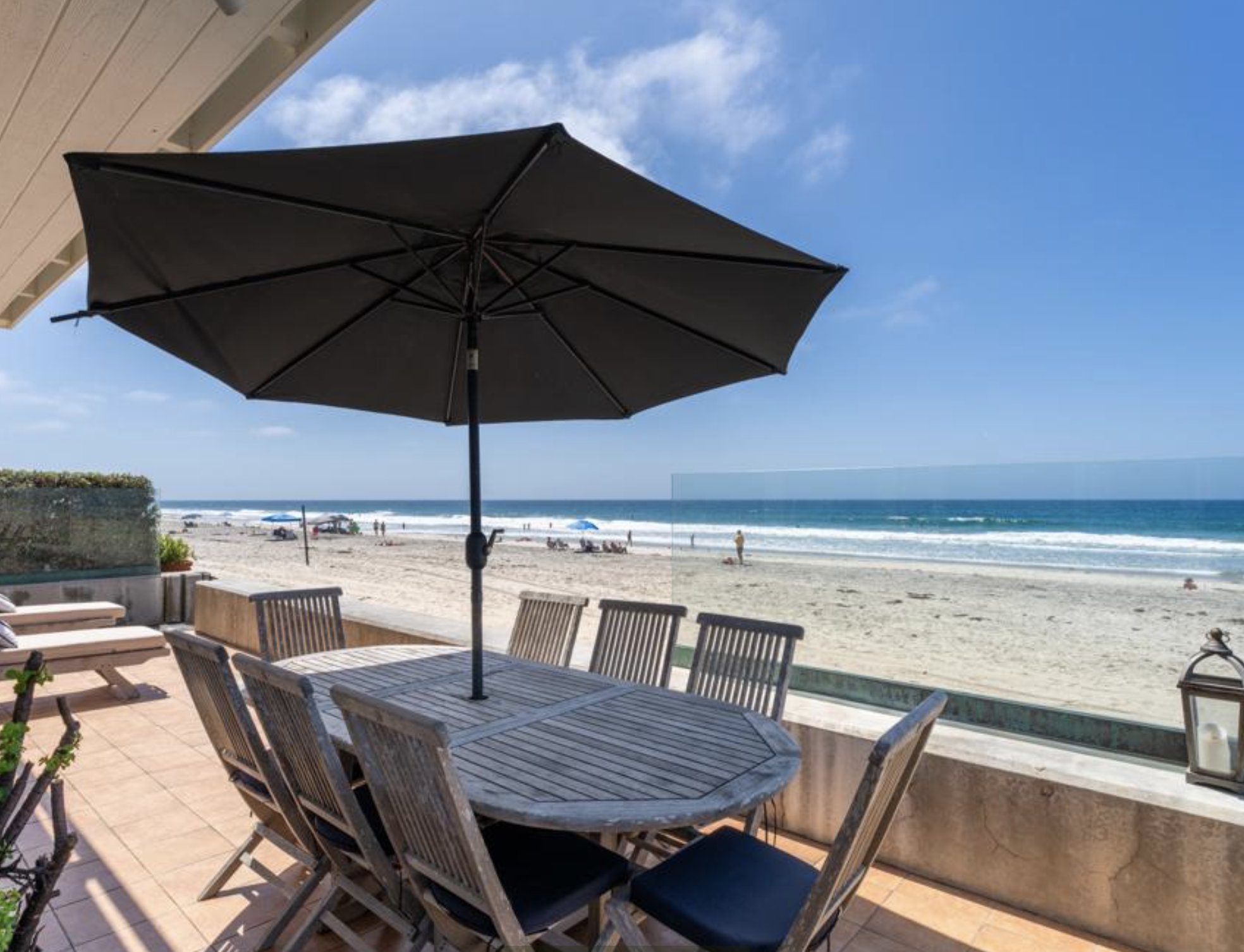 Oceanfront patio in Del Mar California with a wooden dining table, black umbrella, and glass railing overlooking the sandy beach