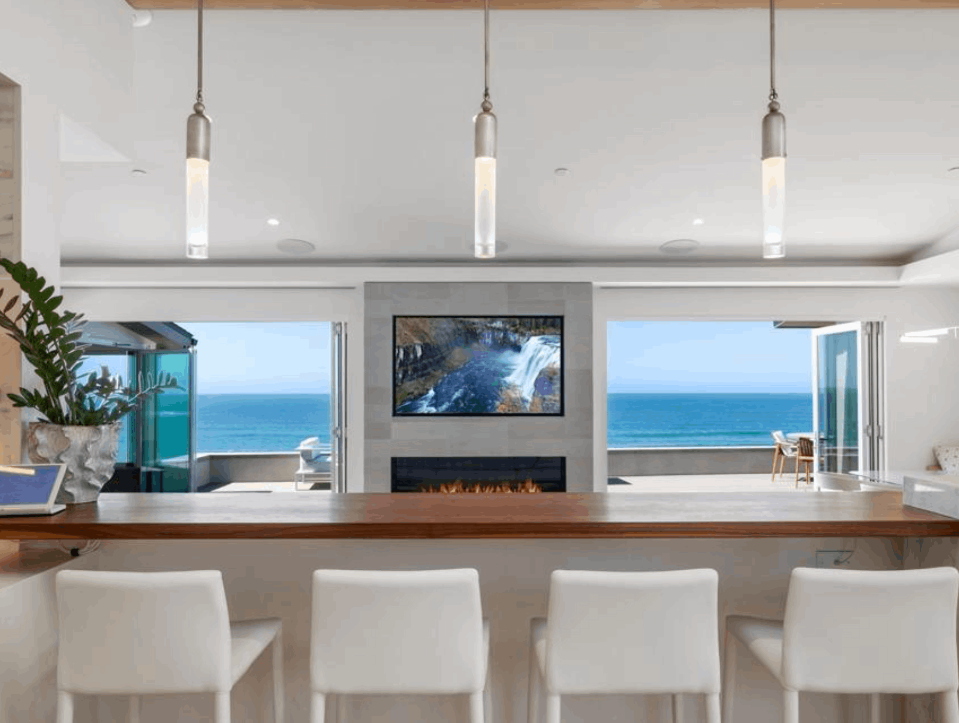 An oceanfront kitchen bar with white stools overlooking a modern fireplace, TV, and panoramic Pacific Ocean views through large glass bifold doors