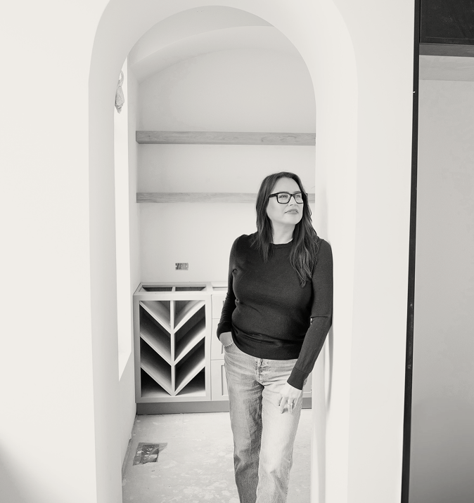 Black and white portrait of a woman standing in an arched doorway in a construction or remodel space, vertical composition showing head to mid-thigh, neutral tones.