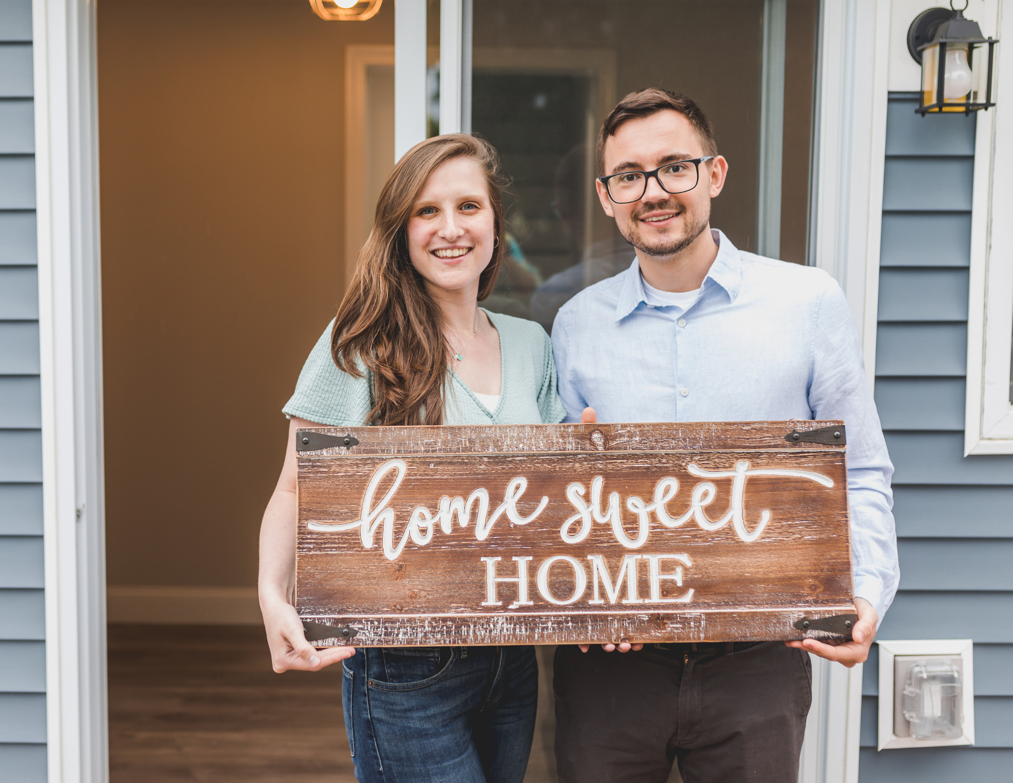 "Smiling couple standing in front of their new home holding a rustic wooden sign that says 'Home Sweet Home'."