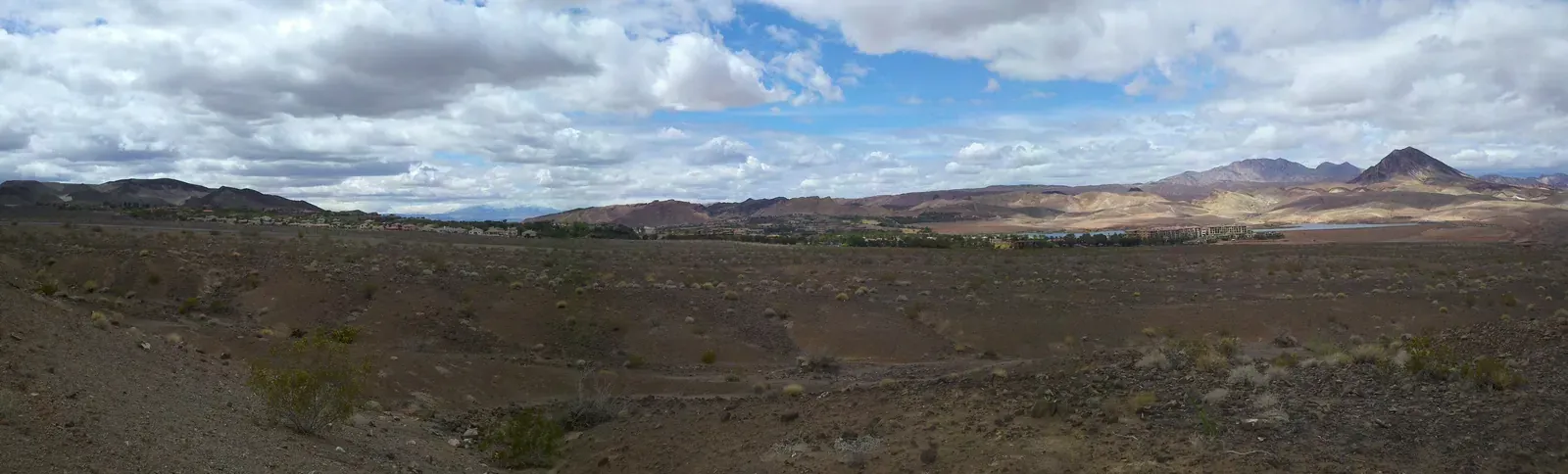 Desert scrub and hills with Lake Las Vegas and its buildings in the distance under a cloudy sky