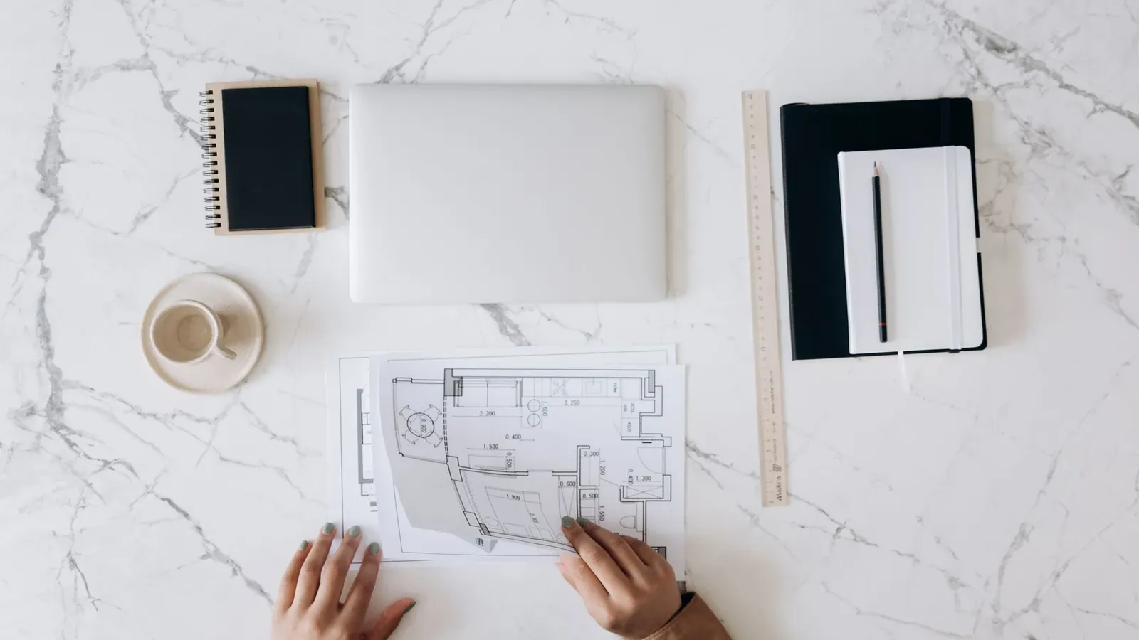Two hands review a printed floor plan on a table with notebooks, a pen and coffee