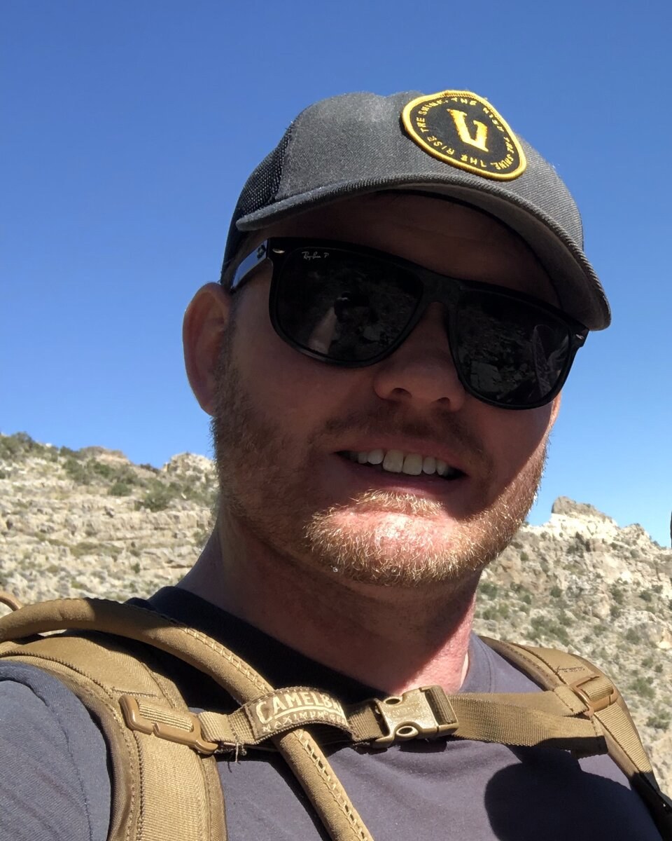 Man on a desert hiking trail wearing a cap and sunglasses, rocky hills under clear sky