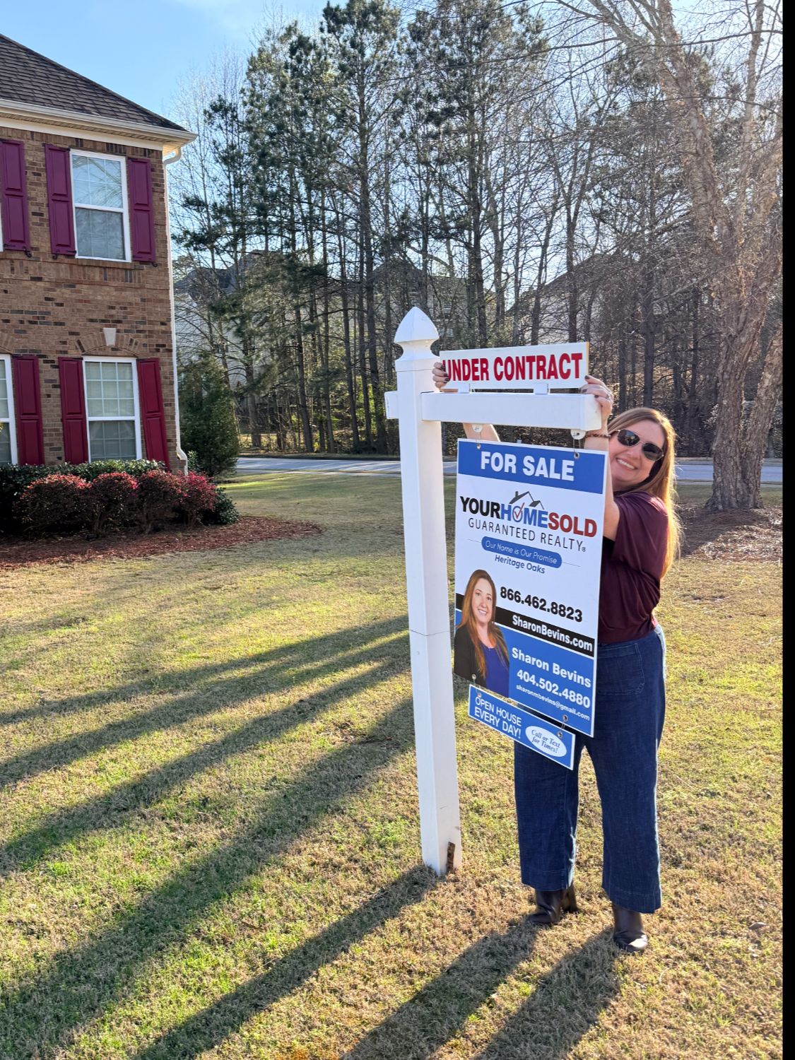 Sharon Bevins, Realtor standing confidently next to a 'Your Home Sold Guaranteed' sign with an 'Under Contract' rider, in front of a large brick residential home.