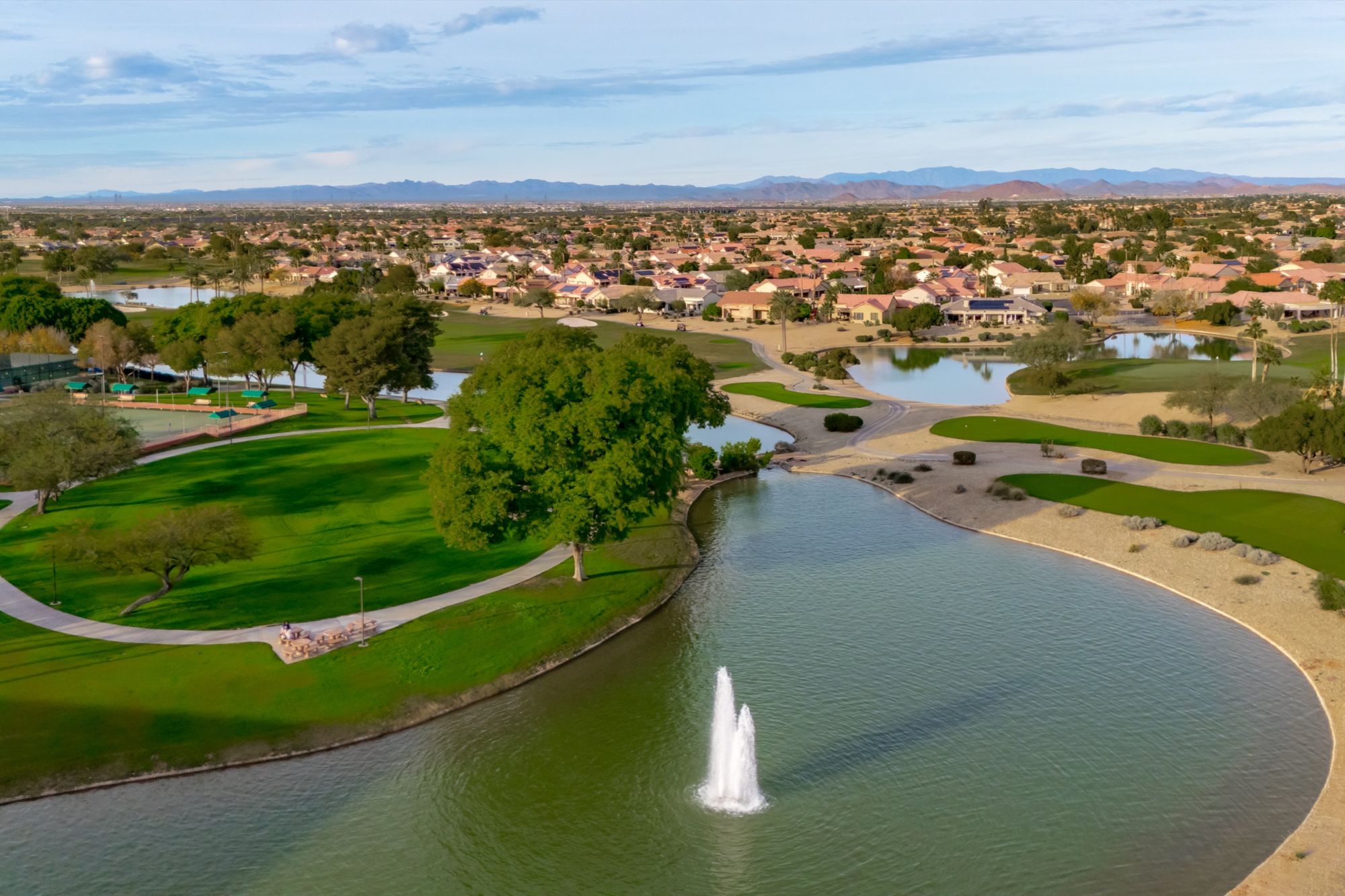 Aerial view of Sun City Grand in Surprise, Arizona, featuring championship golf course fairways, scenic lakes with water fountains, walking paths, and surrounding homes in one of the West Valley’s most desirable 55+ active-adult communities.