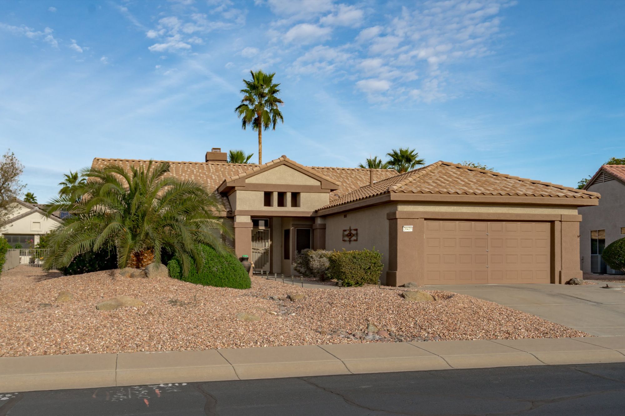 Front exterior of a Cholla model home in Sun City Grand, Surprise AZ, featuring pristine desert landscaping with mature saguaro cacti, a two-car garage, tile roof, and a peaceful 55+ community setting at sunset.