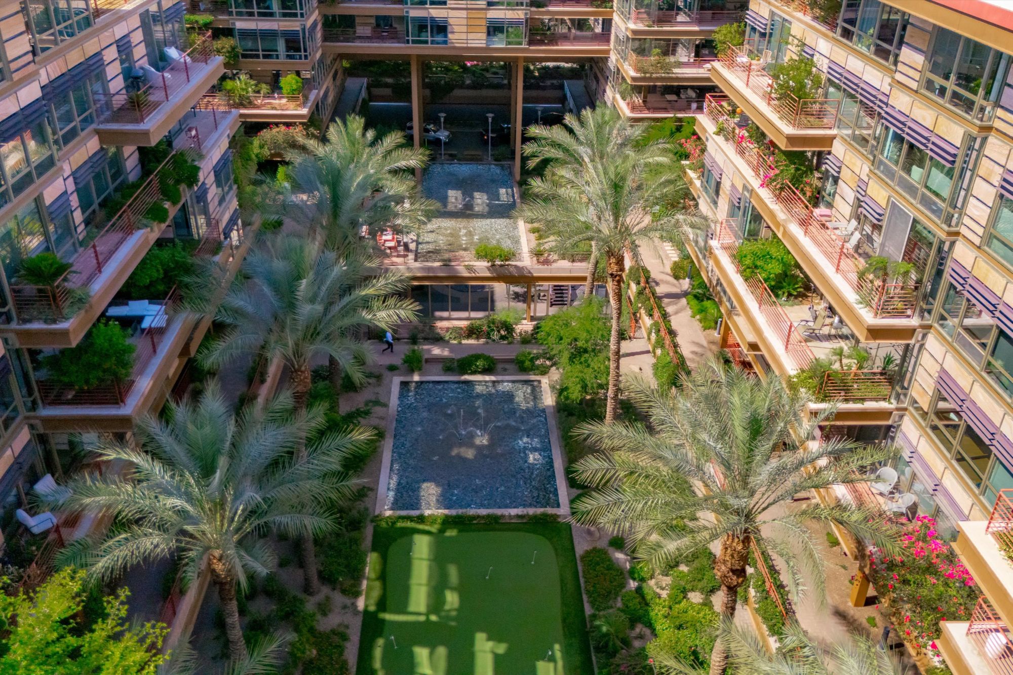 Lush interior courtyard at Optima Camelview Village in Scottsdale, AZ, featuring palm trees, water features, putting green, and private condo balconies. This luxury condo community near Scottsdale Fashion Square offers a tranquil resort-style setting with mid-term and long-term rental options, walkable access to Old Town, and premium amenities including pools, fitness center, concierge, and on-site dining.