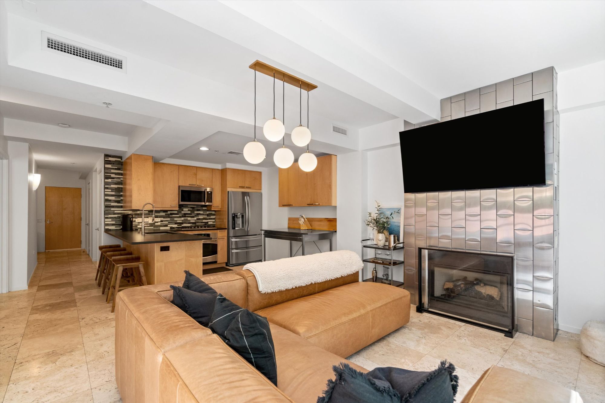 Open-concept living room and kitchen in a remodeled Optima Camelview Village condo in Scottsdale, AZ. Features include a leather sectional, modern fireplace with custom tile surround, pendant lighting, and a chef-style kitchen with Viking appliances, quartz counters, and designer backsplash. Located near Scottsdale Fashion Square, this luxury condo offers rental flexibility and resort-style amenities.