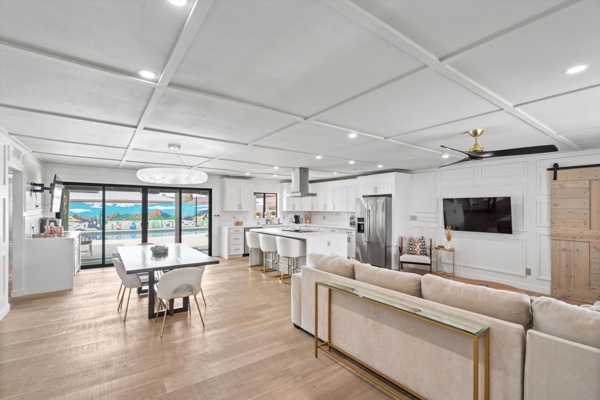 Bright and modern open-concept interior of a Scottsdale vacation rental featuring a designer kitchen with white cabinetry, quartz countertops, stainless steel appliances, spacious island with gold-accented barstools, elegant dining area, and cozy living room—all overlooking the backyard pool and mural through large sliding glass doors.