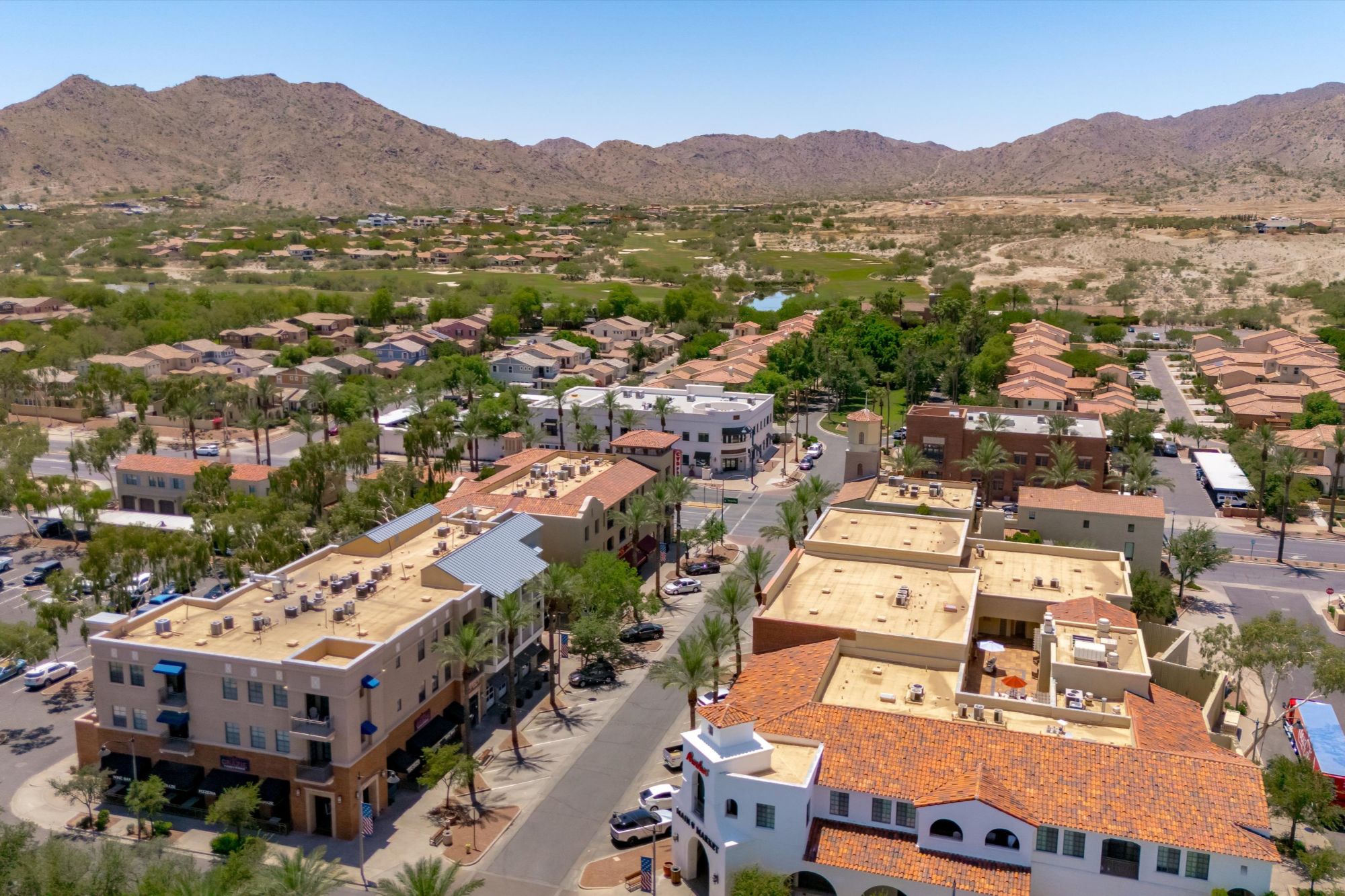 Aerial view of Verrado Main Street in Buckeye, Arizona showcasing the walkable town center, palm-lined streets, local shops, restaurants, and nearby residential neighborhoods set against the White Tank Mountains in the Verrado master-planned community.