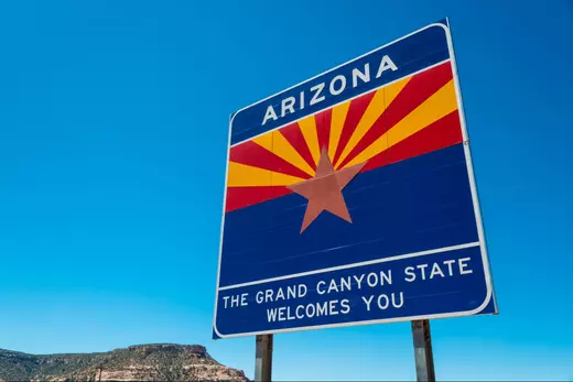 Arizona welcome sign with the state flag and ‘The Grand Canyon State Welcomes You’ message under a bright blue sky, representing the experience of moving to Arizona and why new residents are choosing the state.