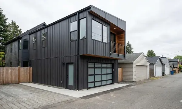 Modern two-story DADU built over a two-car garage on a Seattle alleyway, featuring dark vertical metal siding, a modern glass-panel garage door and a private balcony.