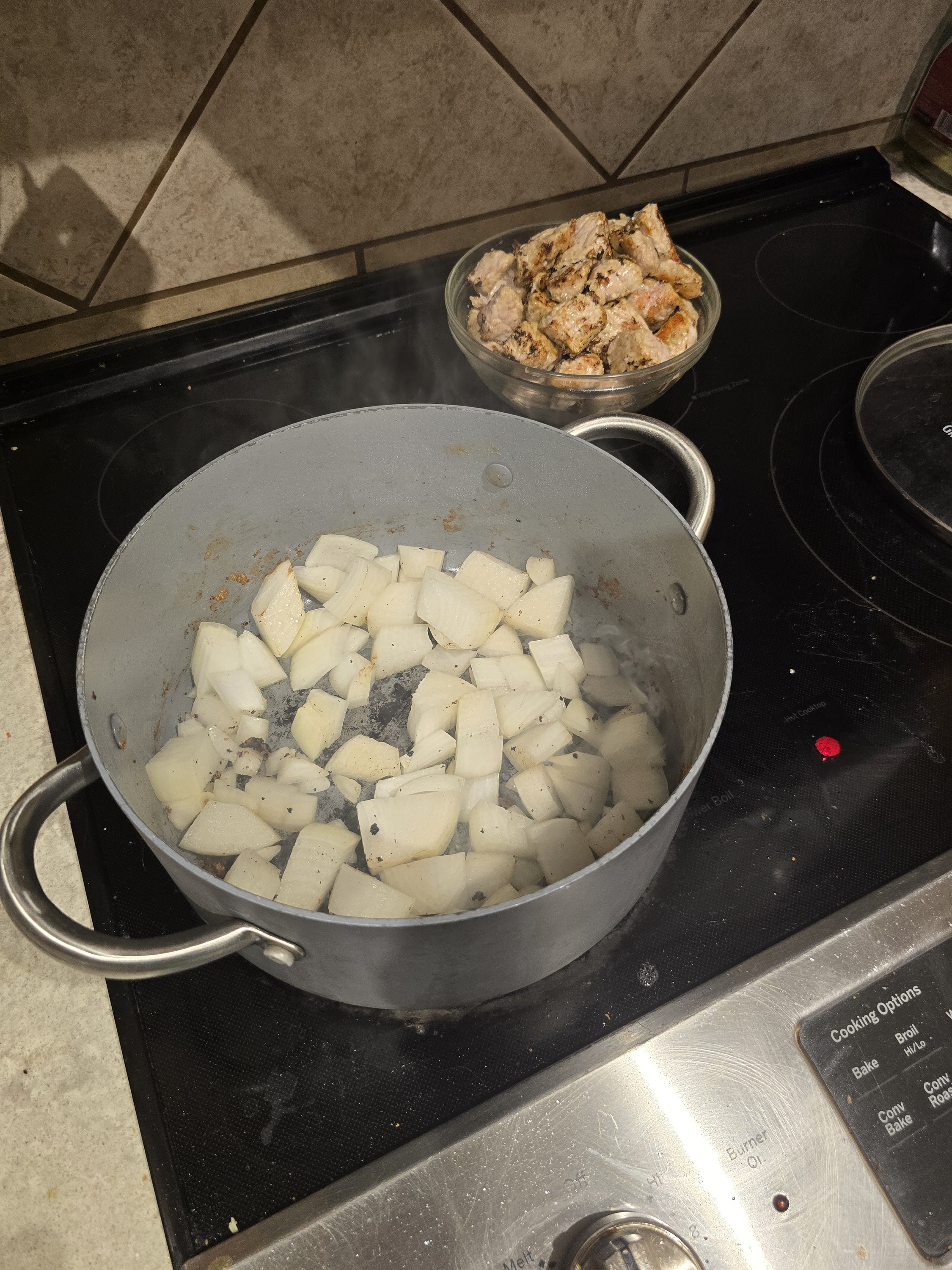 A large silver pot on the stovetop with diced onions simmering in the early stages of cooking. In the background, a bowl of browned pork pieces sits ready to be added to the stew.