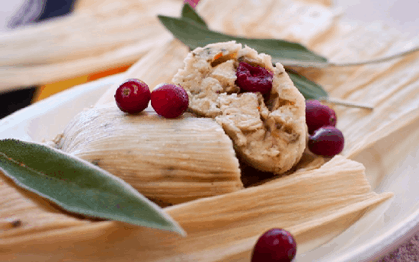 Close-up of a tamale on a plate, filled with turkey and cranberries, garnished with fresh cranberries and sage leaves.