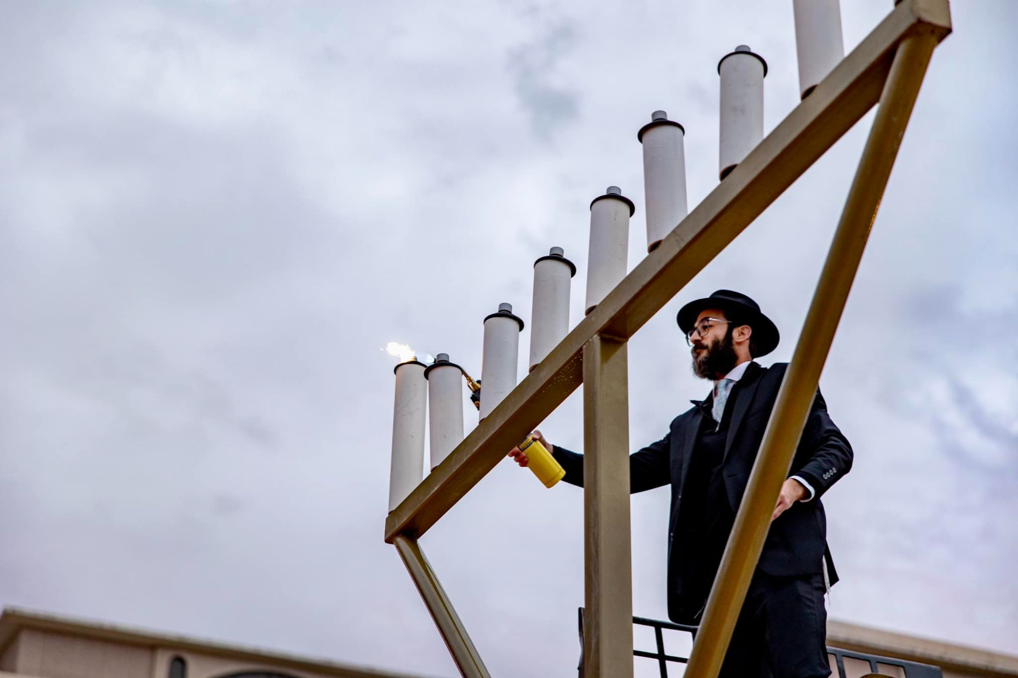Man in a black suit, tie, and hat lights a giant outdoor menorah with a torch against a cloudy sky.