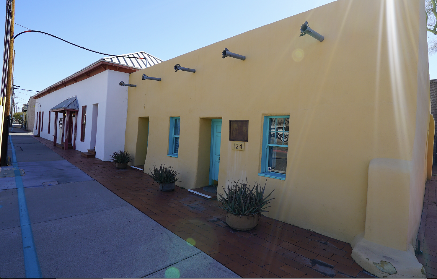 Street-level view of a historic adobe building with light yellow and white stucco walls, turquoise doors and windows, and potted agave plants along the walkway.