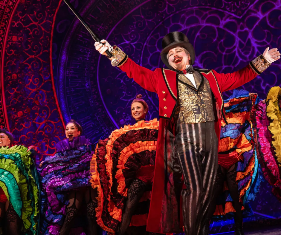 Theatrical scene with a performer in a red tailcoat and top hat leading dancers in colorful can-can skirts on a richly decorated stage.