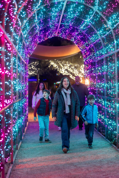 A family walking through a tunnel of colorful holiday lights at Tucson Botanical Gardens during the LightsUp! winter evening event.