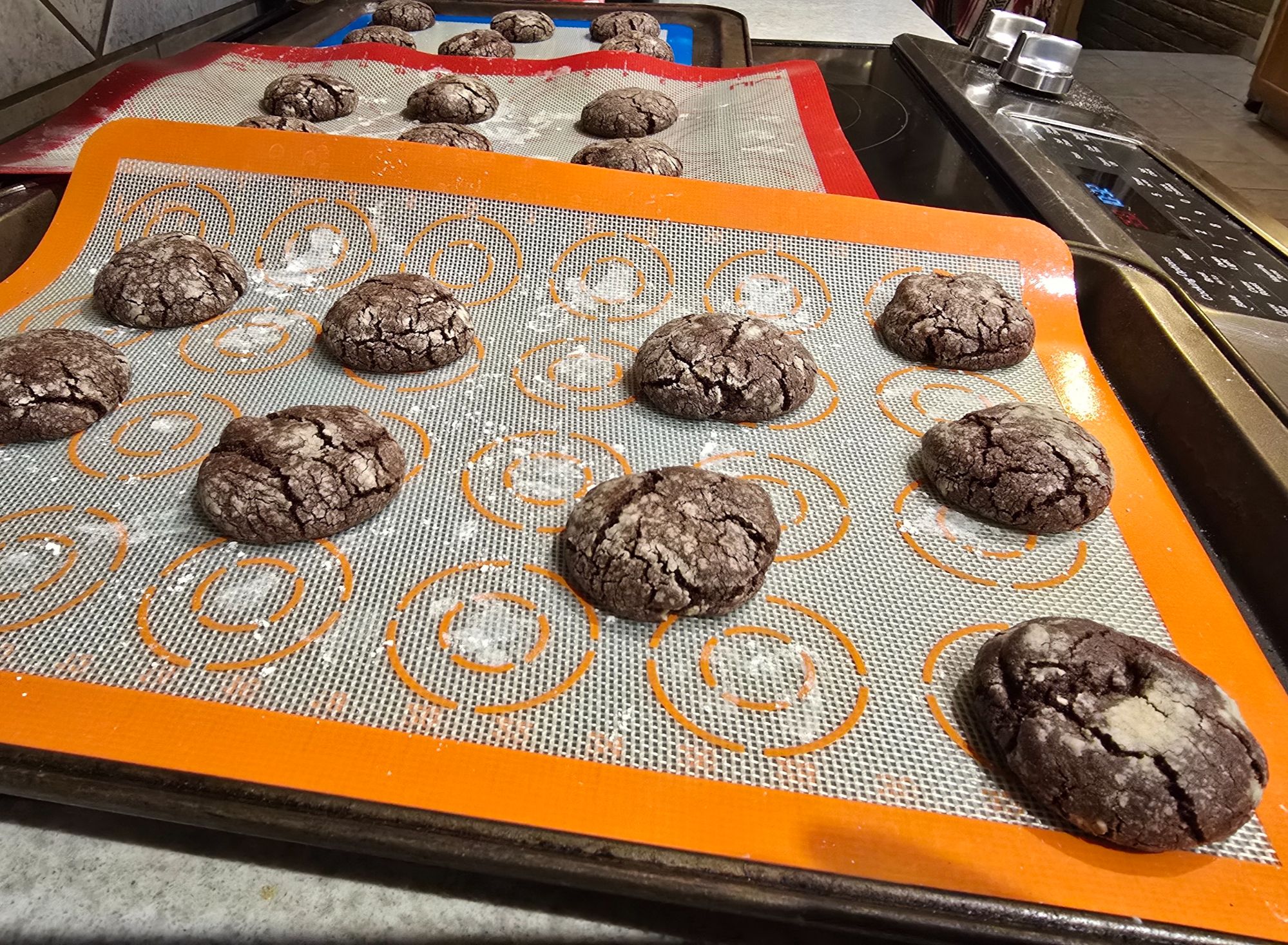 Freshly baked Mexican chocolate crinkle cookies cooling on orange, red, and blue silicone baking mats. The cookies have classic crackled tops and rich, dark chocolate color.