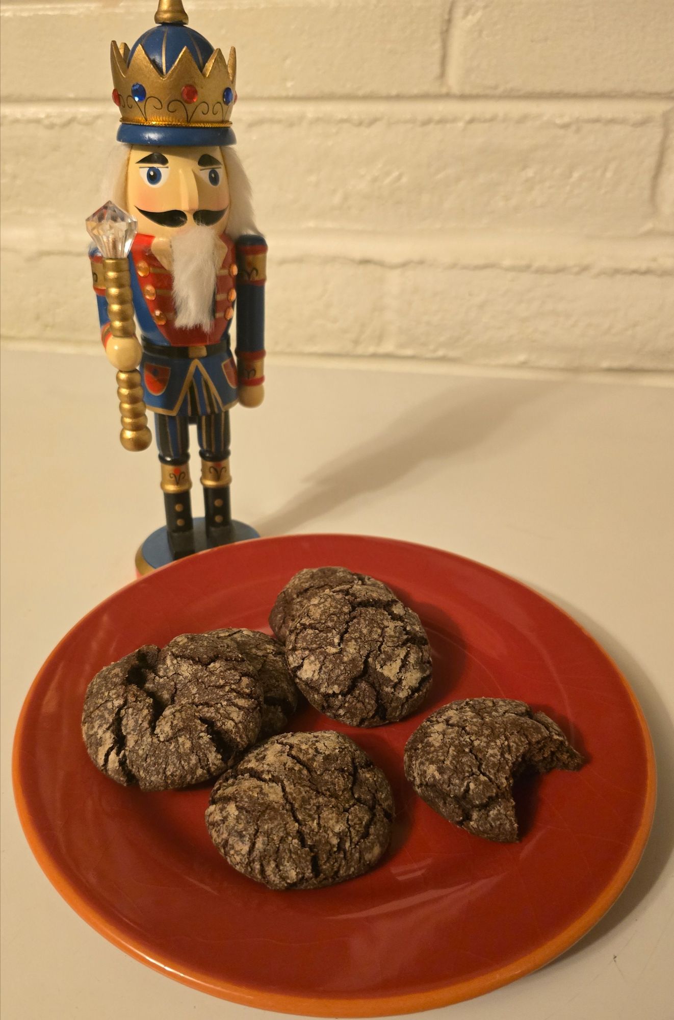 A red plate of finished Mexican chocolate crinkle cookies with crackled powdered-sugar tops sits beside a decorative blue and gold nutcracker figurine against a white brick background. One cookie has a bite taken out.