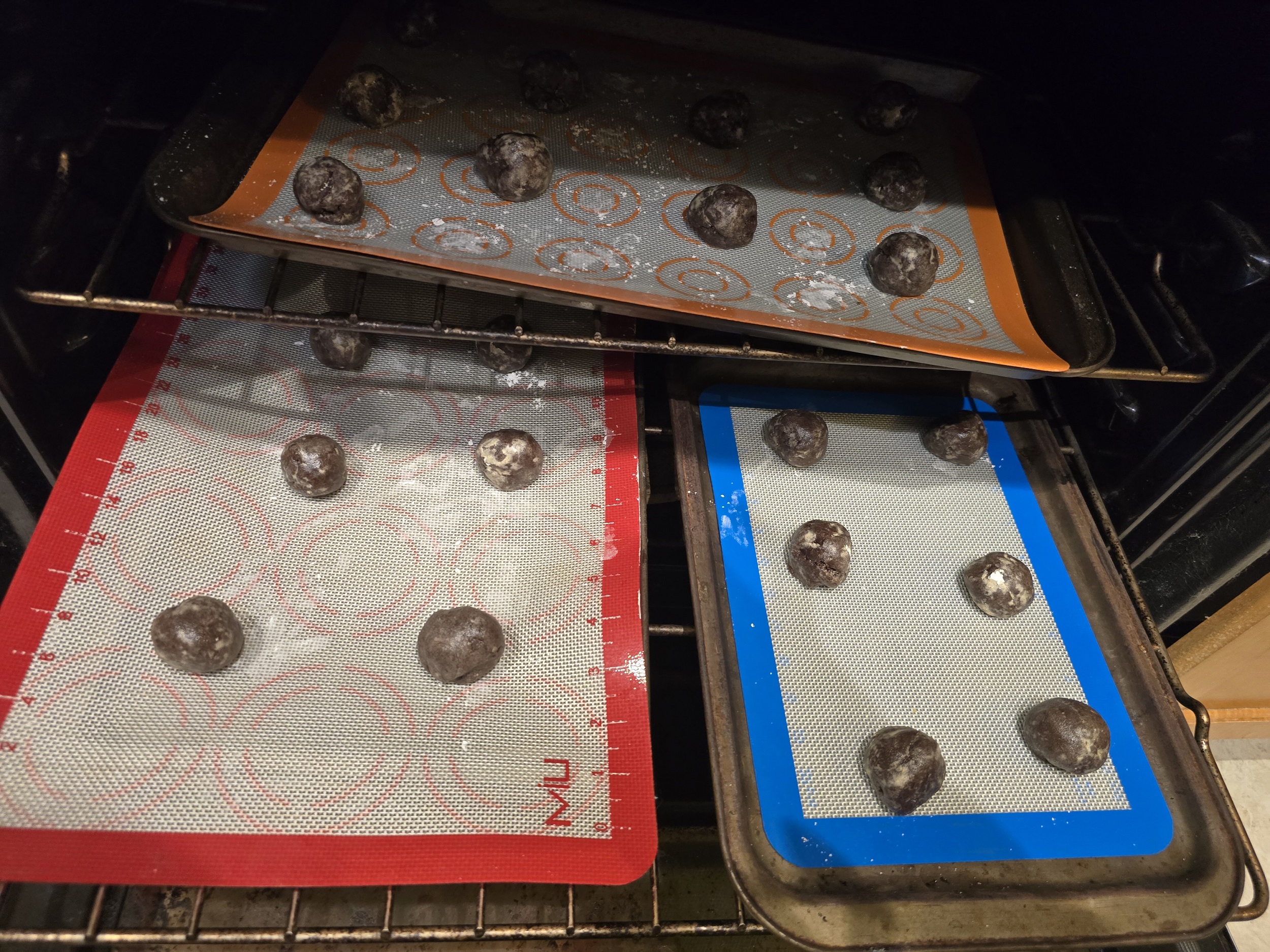 Cookie dough balls coated lightly in powdered sugar baking in the oven on three sheet pans lined with colorful silicone mats. The round chocolate dough balls are spaced evenly as they begin to puff and crackle during baking.