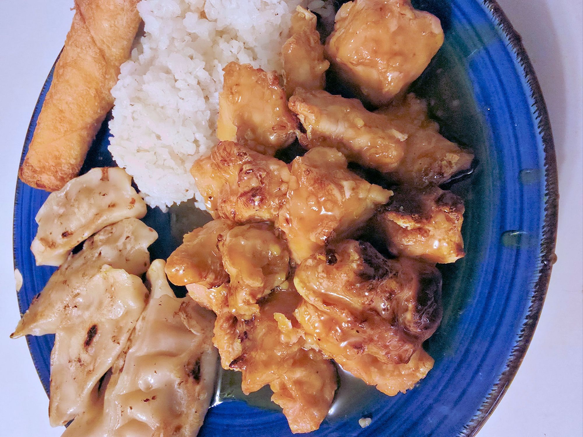 Plate of Chinese-style lemon chicken served with white rice, vegetable dumplings, and a crispy egg roll, with glossy lemon sauce coating the chicken pieces.
