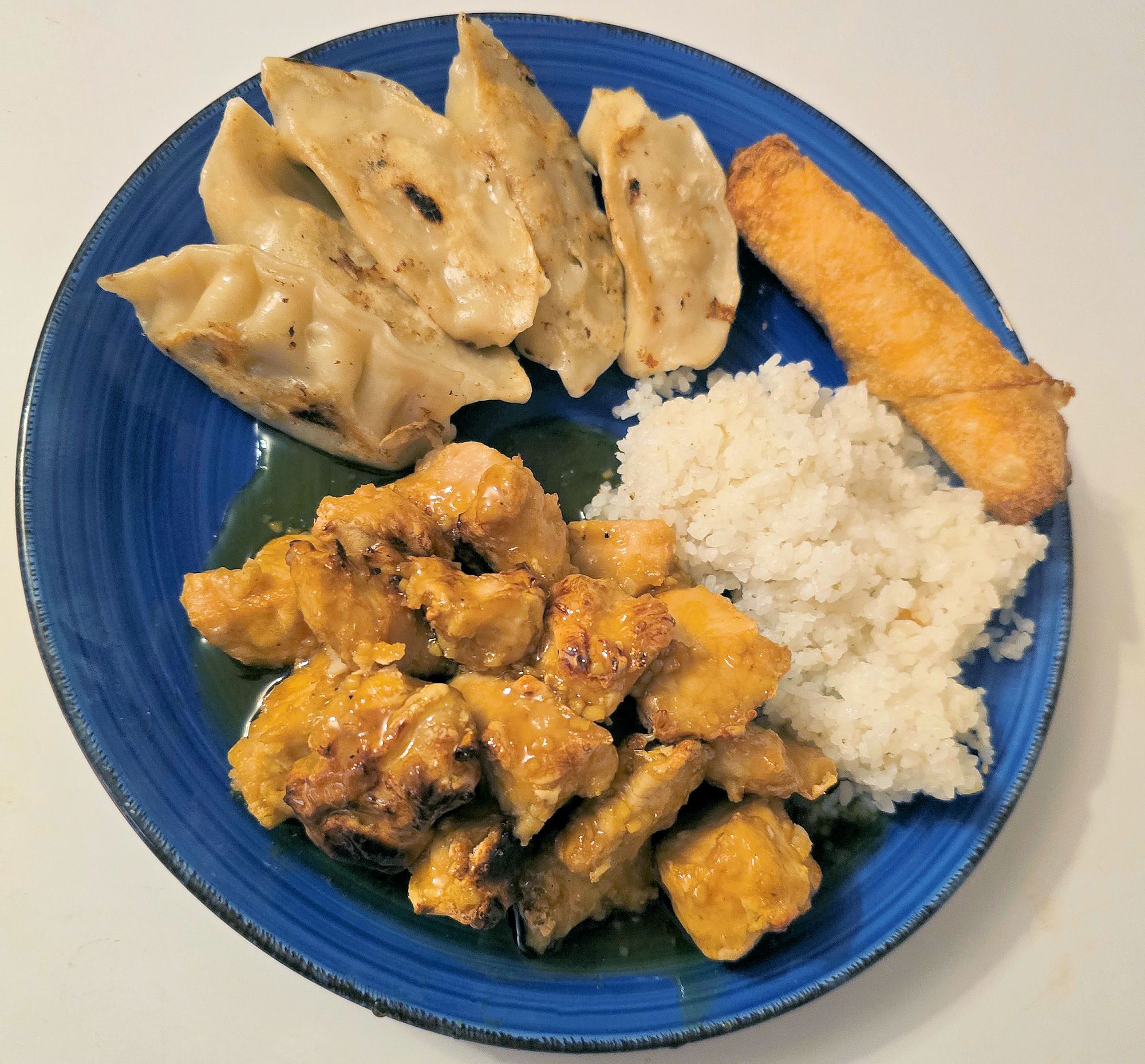 Full plate of air-fried lemon chicken with steamed white rice, pan-seared dumplings, and an egg roll, arranged on a blue ceramic plate.