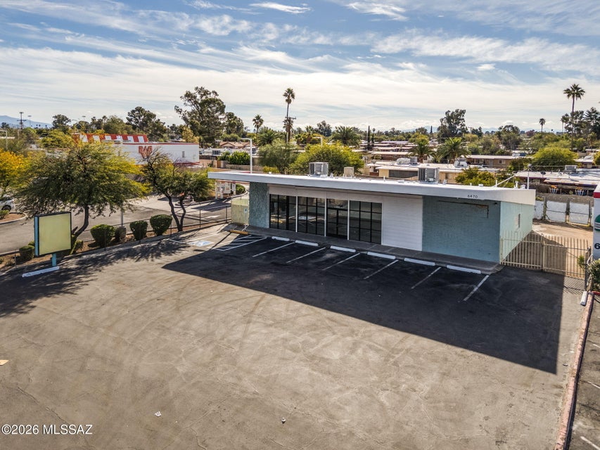 Aerial view of a single-story commercial building with large front windows and a freshly paved parking lot in Tucson, Arizona. The property sits on a corner lot with surrounding trees, nearby businesses, and mountain views in the distance under a partly cloudy sky.