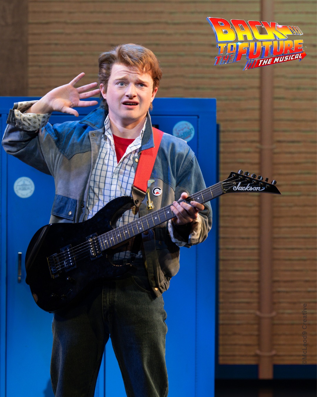 Promotional photo for Back to the Future: The Musical. A young male actor in 1980s costume — denim jacket, plaid shirt, red vest — plays a black Jackson electric guitar in front of blue school lockers.