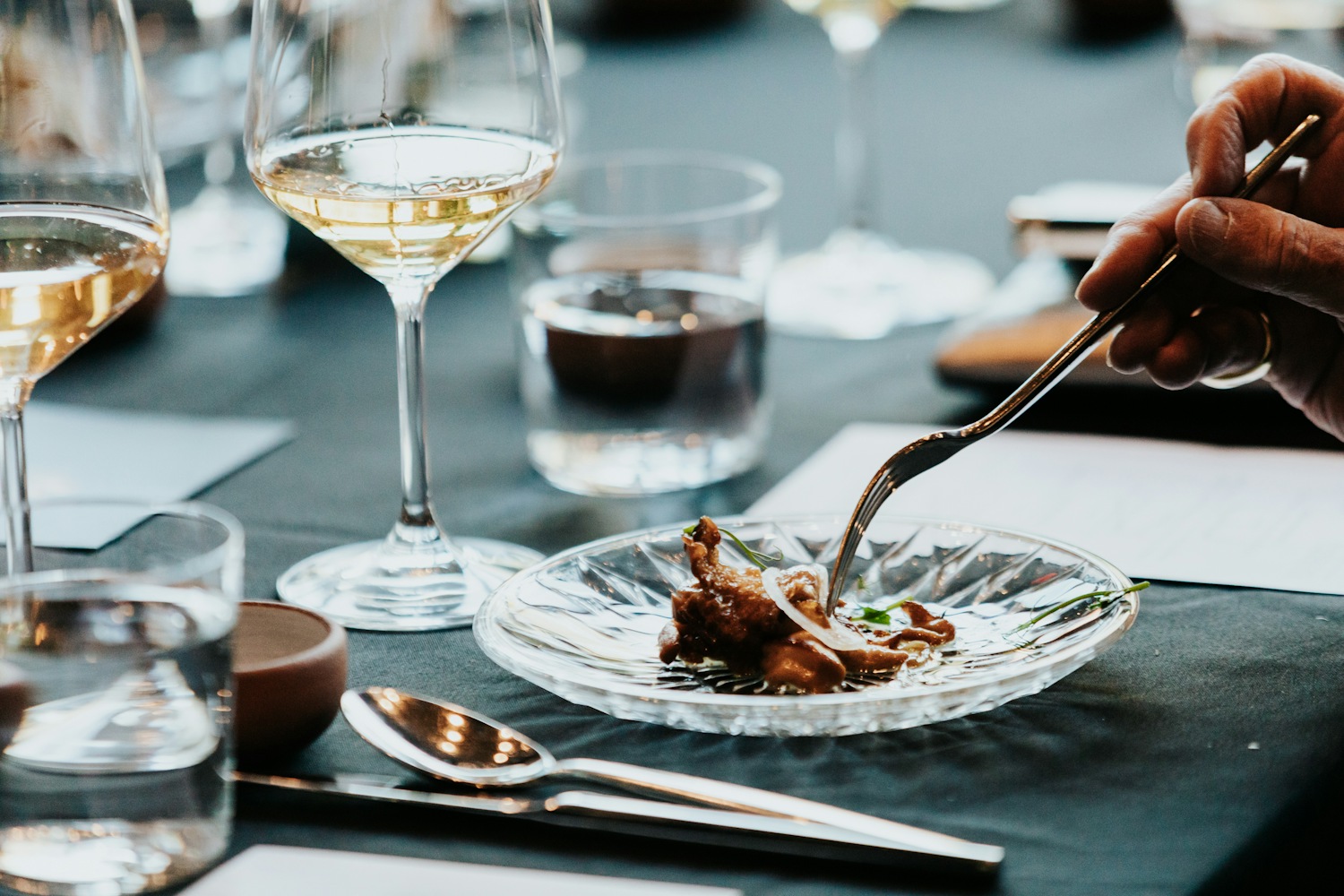 Close-up of a fine dining place setting: a crystal plate with a small portion of braised meat and herbs, a white wine glass, water glass, and silverware on a dark tablecloth. A hand holds a fork over the plate.