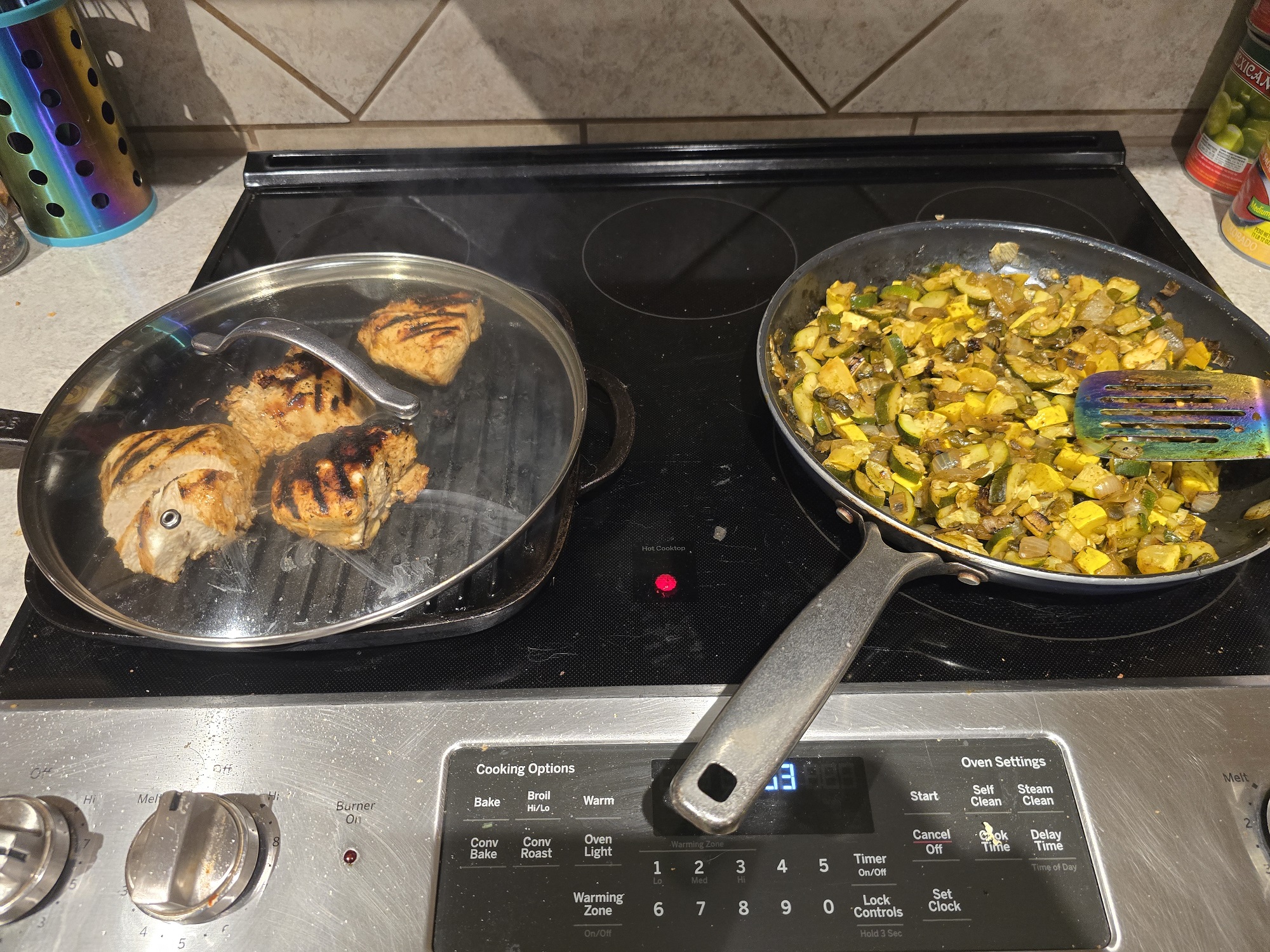 A stovetop with two pans cooking simultaneously. The left pan is a ridged grill pan with three bone-in chicken thighs showing grill marks. The right pan contains sautéed zucchini and yellow squash with onion, being stirred with a rainbow-handled spatula.
