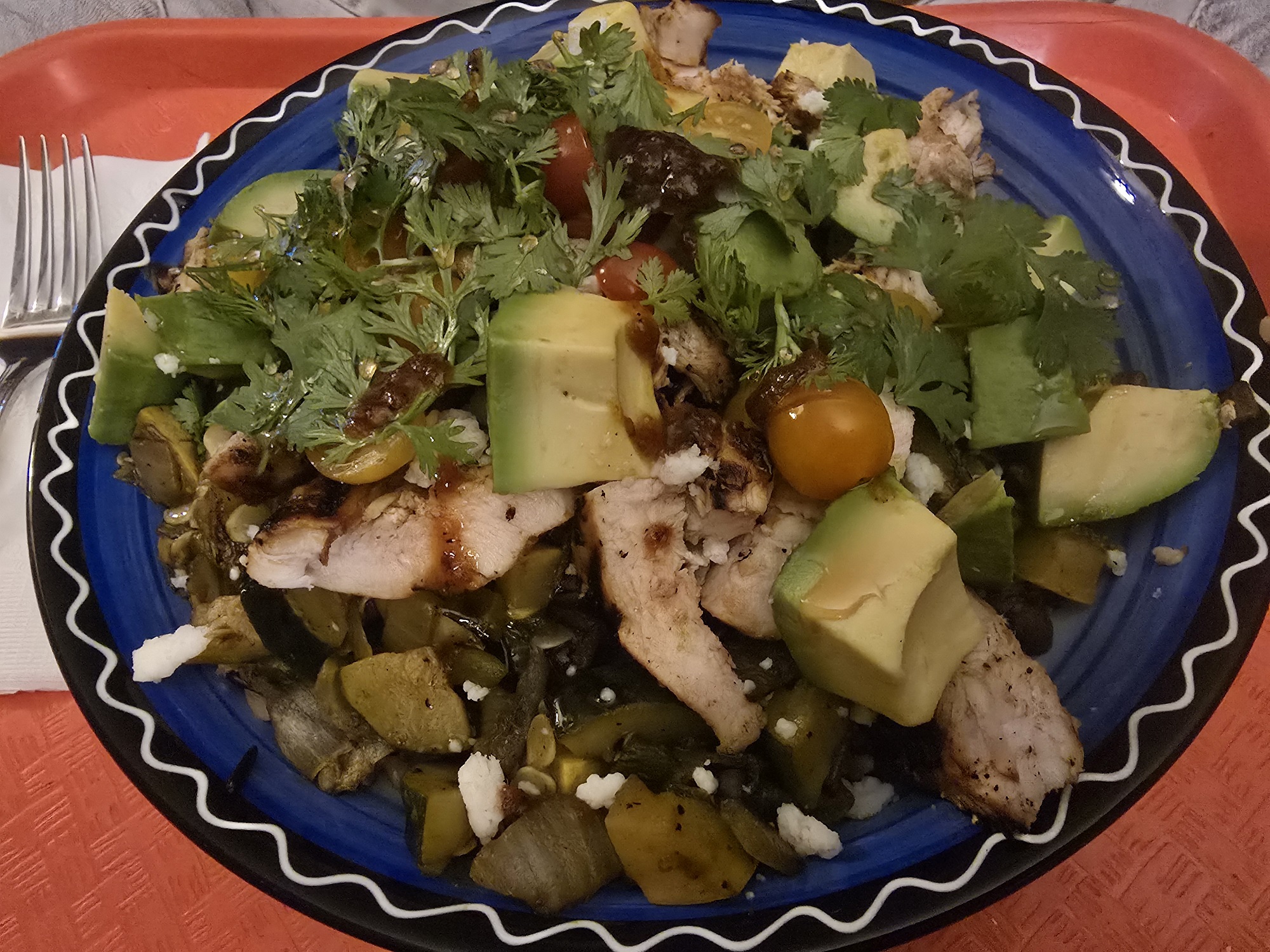 A finished plated meal on a blue and black Talavera-style ceramic plate, served on an orange tray. The dish features sliced grilled chicken, sautéed zucchini and squash, chunked avocado, halved cherry tomatoes, crumbled cotija cheese, fresh cilantro, and what appears to be a dark sauce drizzled over the top.