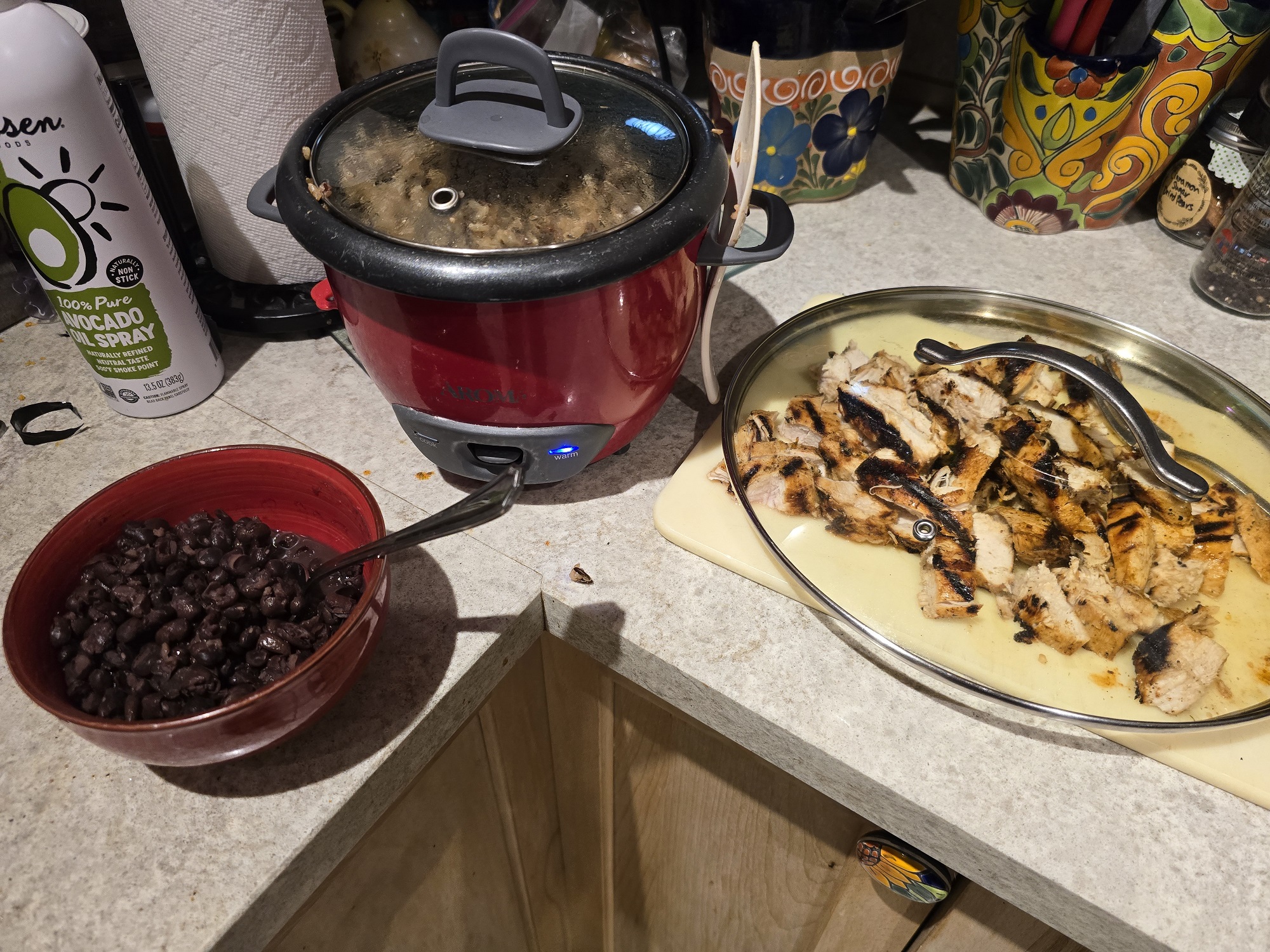 A kitchen counter with three components of a meal in progress: a red rice cooker with a glass lid containing cooked rice, a red ceramic bowl filled with black beans, and a silver oval platter with sliced grilled chicken thighs showing char marks. A can of avocado oil spray is visible on the left.