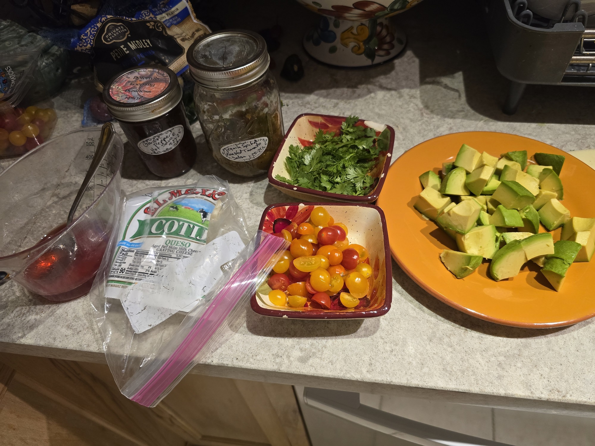A kitchen counter with meal toppings laid out: a bag of El Mexicano cotija queso, two small square bowls containing fresh cilantro and halved multicolored cherry tomatoes, a plate of chunked avocado, and two mason jars in the background labeled as homemade sauces or dressings. A bag of petite medley tomatoes is visible in the upper left.